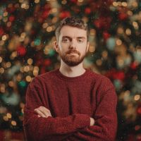 Bearded man with arms crossed in a maroon sweater standing before blurred Christmas tree lights.