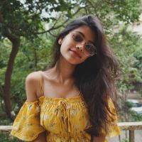 Smiling young woman with long dark hair wearing an off-shoulder yellow floral top and round sunglasses on one eye, outdoors with trees.