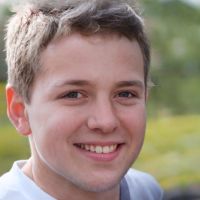 Young smiling man with short brown hair looking at the camera against a blurred green outdoor background.
