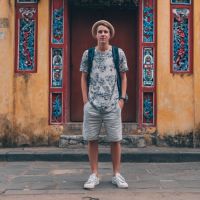 Young man wearing a floral T-shirt, shorts and backpack before a red doorway in a weathered yellow wall with colorful tiles.