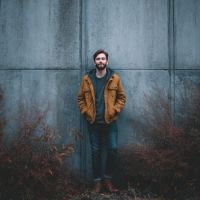 Bearded man in a brown jacket and jeans stands with hands in pockets against a concrete wall among sparse reddish shrubs.
