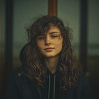 Woman with wind-tousled curly brown hair and neutral expression wearing a dark jacket against a blurred outdoor wooden post.