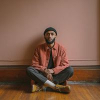 Man wearing a beanie and salmon jacket sits cross-legged on a wooden floor against a pale pink wall, hands resting on knees.