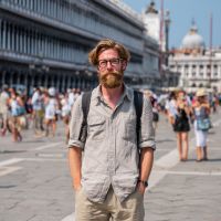 Bearded man with glasses in a light shirt and backpack standing in a crowded sunny plaza with a domed church behind him.