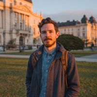 Bearded man in denim shirt and navy jacket with backpack standing on grass before sunlit historic buildings.