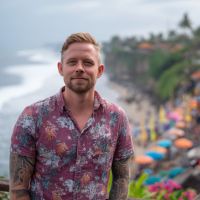 Man with tattoos in a pink floral shirt standing on a cliff overlooking a crowded beach with umbrellas on Penida, Bali.