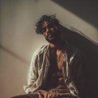 Man with curly hair and beard wearing an open light shirt and necklace, sitting against a wall in dramatic shadowed light.