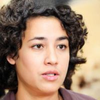 Person with short curly dark hair and a neutral expression, blurred indoor background.