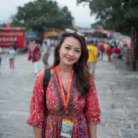 Smiling woman in a red patterned dress with sunglasses on her head and a lanyard badge, standing on a crowded stone walkway.