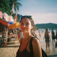Young woman smiling at camera on a crowded tropical beach with sunglasses on her head, palm trees and vendors behind her.
