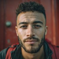 Close-up of a bearded man with short curly hair and freckles, wearing backpack straps in front of a red wooden door.