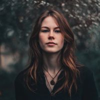 Woman with long wavy auburn hair, wearing a dark top and pendant necklace, standing before a blurred leafy background.