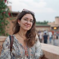 Woman with brown hair and glasses, sunglasses on her head, wearing a patterned top by a ledge with blurred crowd behind.