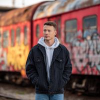 Man in a black denim jacket and gray hoodie standing before graffiti-covered red train cars, hands in pockets, looking sideways