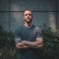 Bearded man with short hair and forearm tattoos standing with arms crossed in a gray T-shirt against a textured stone wall and low plants.
