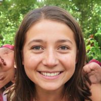 Smiling young woman with shoulder-length brown hair and brown eyes, posing outdoors against a leafy green background.