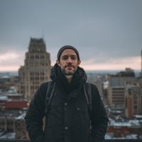 A man wearing a beanie and dark coat with a backpack stands on a rooftop with a city skyline under an overcast sky.
