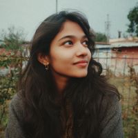 Young woman with long dark hair and hoop earrings gazing upward with a soft smile outdoors near a fence.