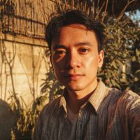 Young man with short dark hair wearing a striped shirt, lit by warm golden-hour sun, with plants and a wooden fence behind him.