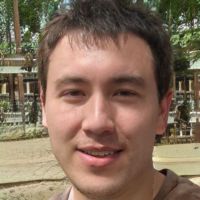 Young man with short dark hair and light stubble smiling outdoors with wooden railings and trees in the background.