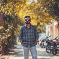 Bearded man wearing sunglasses and a plaid shirt stands with hands in pockets on a tree-lined sidewalk, motorcycles parked behind him.