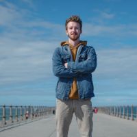 Young man standing on a pier with arms crossed, smiling, wearing a blue jacket over a mustard hoodie under a clear blue sky.