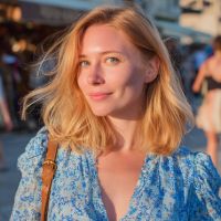 Smiling blonde woman with shoulder-length hair in a blue patterned dress, sunlit at a busy outdoor market.