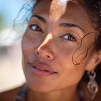 Close-up of a smiling woman with sunlit skin, loose curls, a diagonal shadow on her face, and a dangling earring.