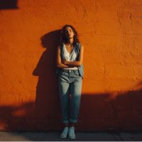 Person leaning against orange wall with arms crossed, wearing denim and white sneakers, long hair and a bold side shadow.