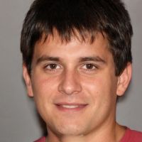 Young man with short dark hair and brown eyes, faint smile, wearing a maroon shirt against a plain gray background.