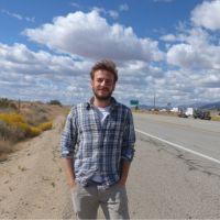 Man in a blue plaid shirt and khaki pants standing with hands in pockets beside a desert highway under a cloudy blue sky.